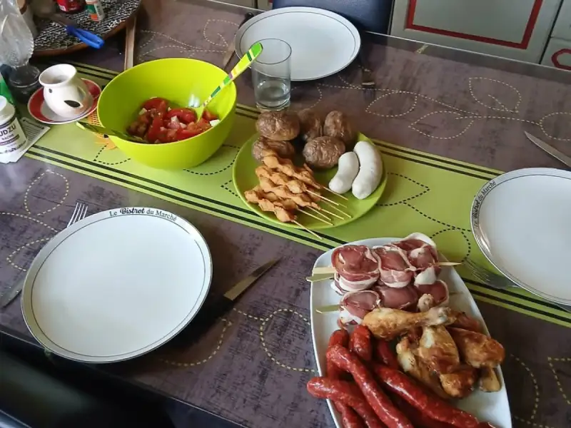 Espetos de frango cru em uma churrasqueira, acompanhados de salsichas, pilão, almôndegas e salada de tomate.