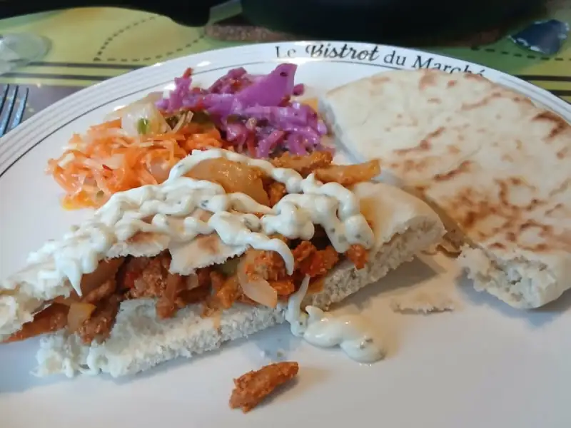 Gyros caseiro, com carne temperada e molho branco com ervas, acompanhado de repolho roxo e cenoura ralada, e uma frigideira ao fundo.
